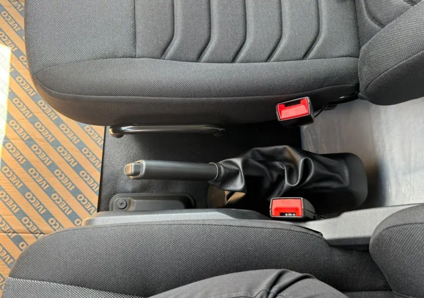 Vue rapprochée de la console centrale entre les sièges noirs du fourgon Iveco Daily, avec frein à main et boucles de ceinture rouges.