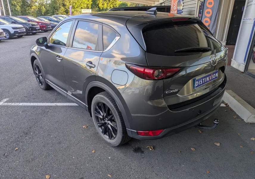 Vue 3/4 arrière droite d'un Mazda CX-5 gris 2020 avec jantes noires et attelage visible sur parking extérieur.
