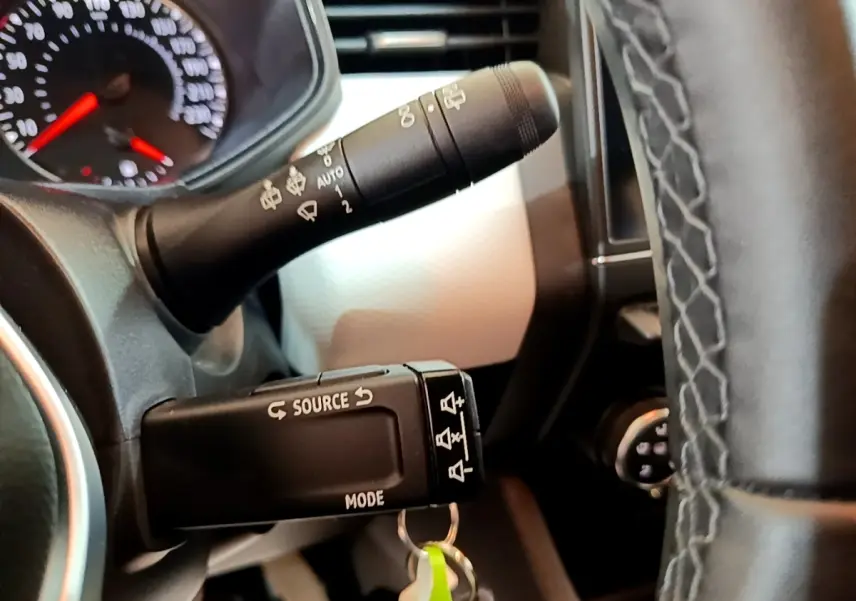 Gros plan sur la commande multimédia au volant cuir noir de la Renault Clio bleu, avec tableau de bord partiellement visible.