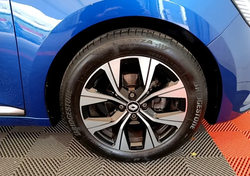 Gros plan sur la roue avant droite d’une Renault Clio bleu 2022 avec jante alliage bicolore et logo Renault central.