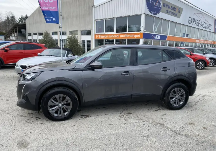 Peugeot 2008 gris foncé vue de profil côté gauche, stationnée devant un garage multimarque.