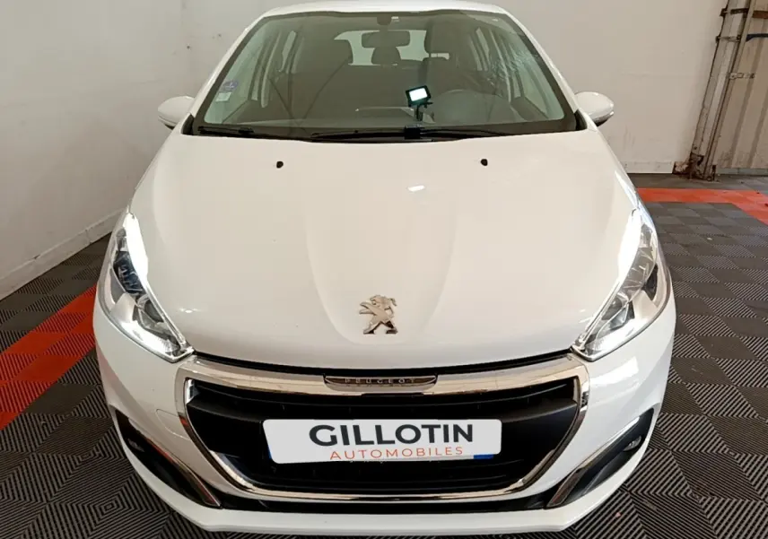 Vue avant d'une Peugeot 208 blanche avec calandre chromée et phares allumés dans un garage.