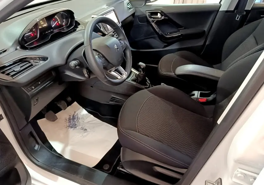 Vue intérieure côté conducteur de la Peugeot 208 blanche, montrant le tableau de bord et les sièges noirs.