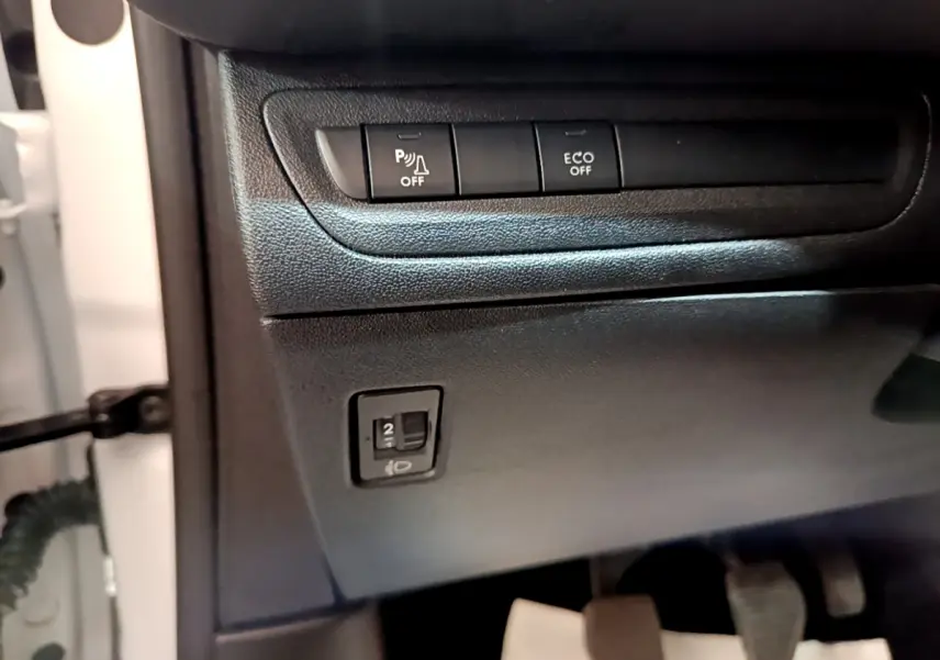 Détail du tableau de bord côté conducteur d'une Peugeot 208 blanche, avec boutons de contrôle et pédales visibles.