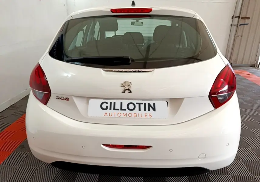 Vue arrière d'une Peugeot 208 blanche stationnée en intérieur, avec feux arrière rouges et logo lion central.