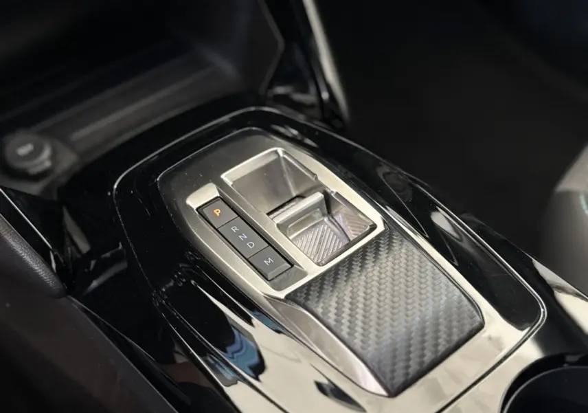 Gros plan sur la console centrale noire brillante avec sélecteur de boîte auto et finition carbone dans un Peugeot 2008 gris clair.