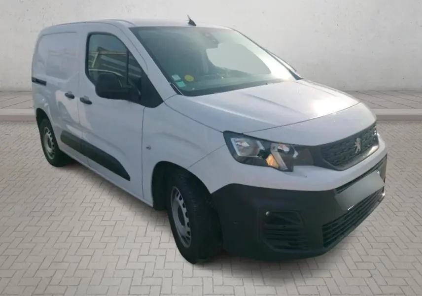 Vue 3/4 avant droite d’un Peugeot Partner blanc 2021 avec pare-chocs noir et calandre Peugeot, stationné sur un sol gravillonné.
