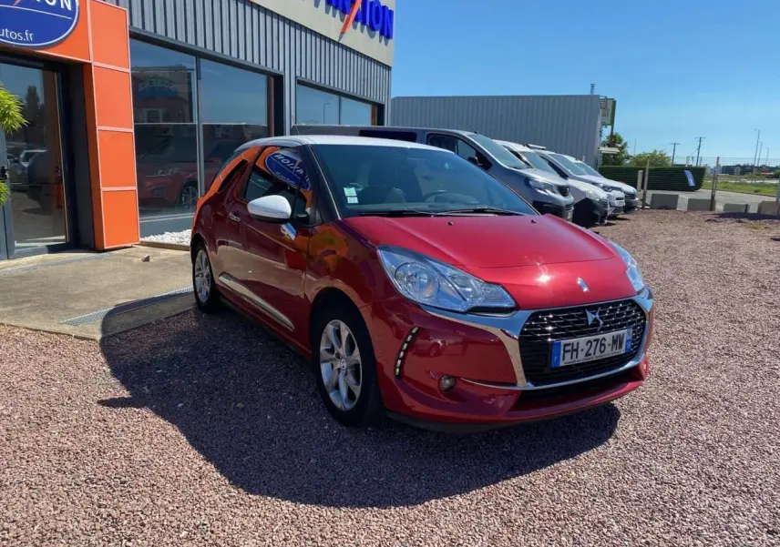DS3 rouge avec toit blanc, vue 3/4 avant droit, garée devant un bâtiment commercial sous un ciel bleu.