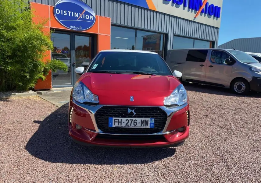 Vue avant d'une DS3 rouge à toit blanc, avec calandre chromée et phares distinctifs, stationnée devant un concessionnaire.