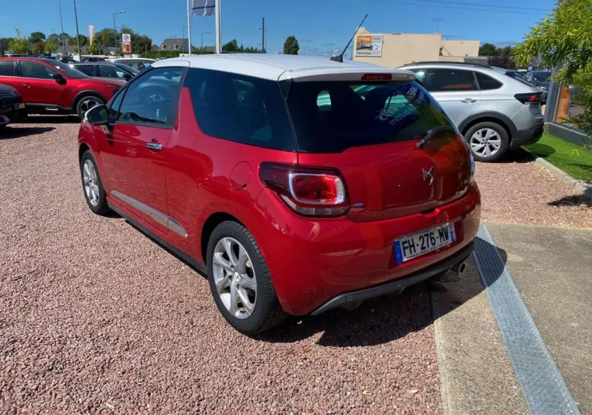 Vue 3/4 arrière droite d'une DS3 rouge à toit blanc, garée sur un sol en gravier sous un ciel dégagé.