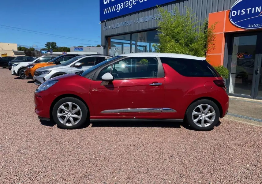 Vue de profil côté gauche d'une DS3 rouge à toit blanc, avec jantes alliage 16 pouces, stationnée devant un garage.
