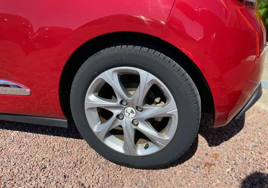 Gros plan sur la roue arrière gauche argentée d'une DS3 rouge avec toit blanc, sur sol gravillonné.