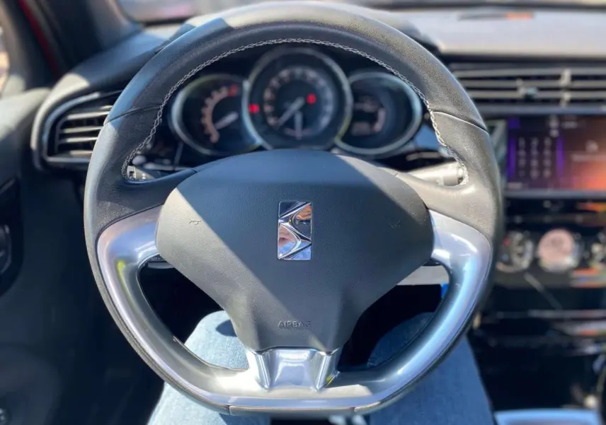 Vue rapprochée du volant DS3 en cuir noir avec logo DS, tableau de bord et écran tactile en arrière-plan flou.