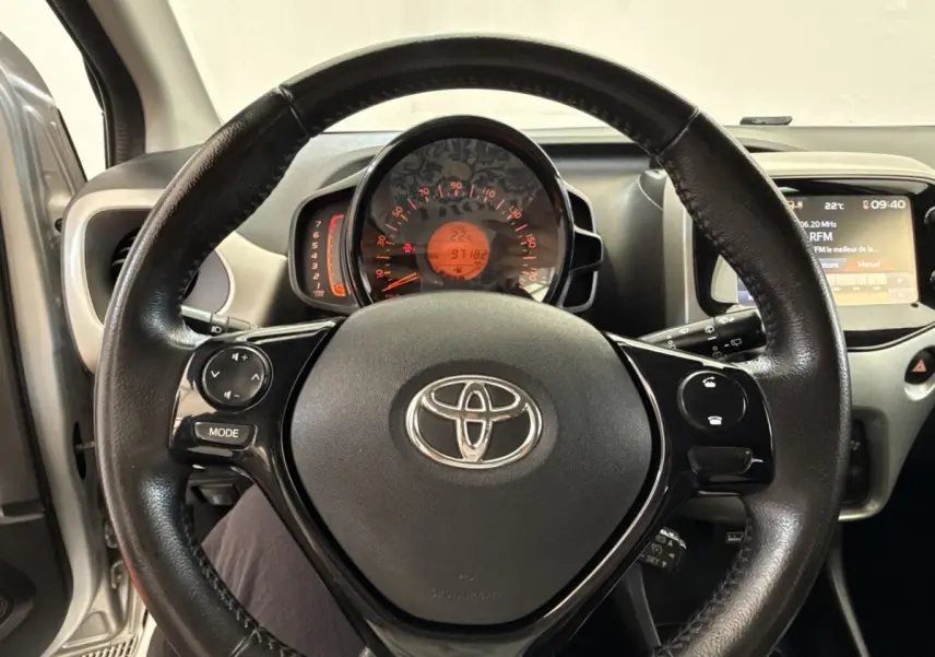 Vue intérieure centrée sur le volant noir de la Toyota Aygo gris clair, avec tableau de bord et écran multimédia visibles.