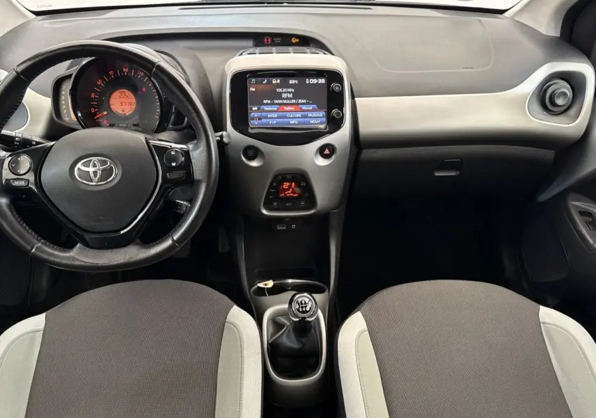 Vue intérieure avant de la Toyota Aygo 2017, montrant le tableau de bord gris clair avec volant, écran tactile et levier de vitesses manuel.