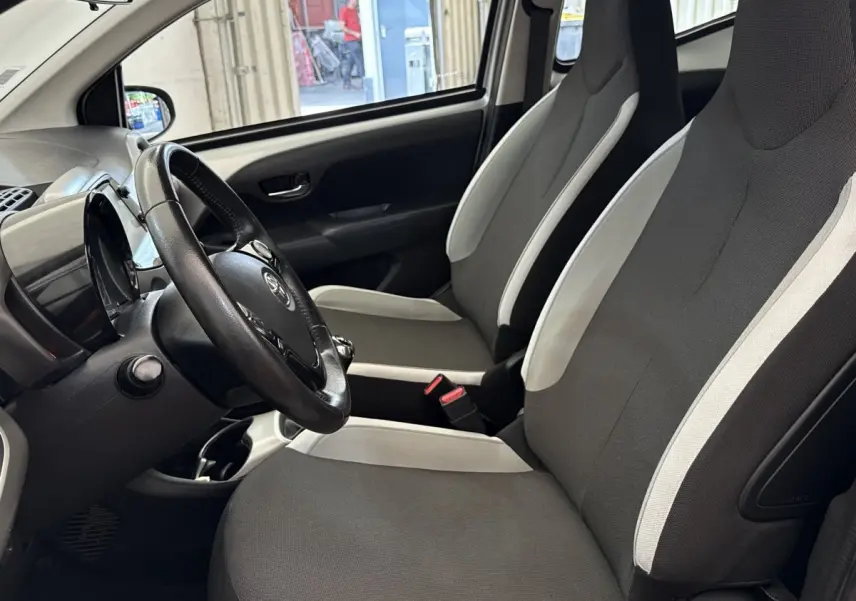 Vue intérieure côté conducteur de la Toyota Aygo gris clair, montrant les sièges avant et le volant noir.