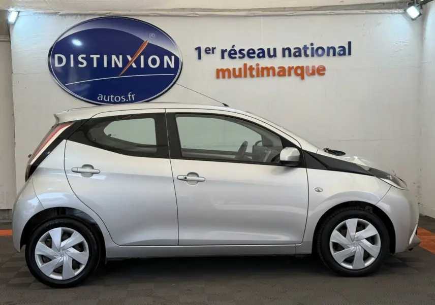 Profil latéral droit d'une Toyota Aygo gris clair 2017, avec jantes et rétroviseurs assortis, en intérieur showroom.