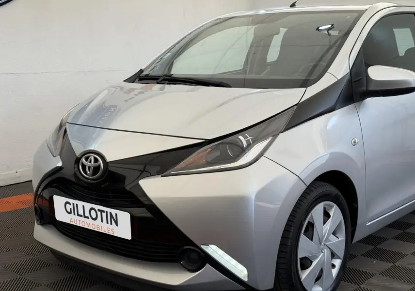 Toyota Aygo gris clair vue 3/4 avant droit, avec calandre noire distinctive et feux LED de jour allumés.