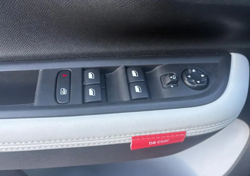 Gros plan sur la commande des vitres et rétroviseurs côté gauche avec surpiqûres blanches et étiquette rouge "be cool" dans une Citroën C3 gris foncé.