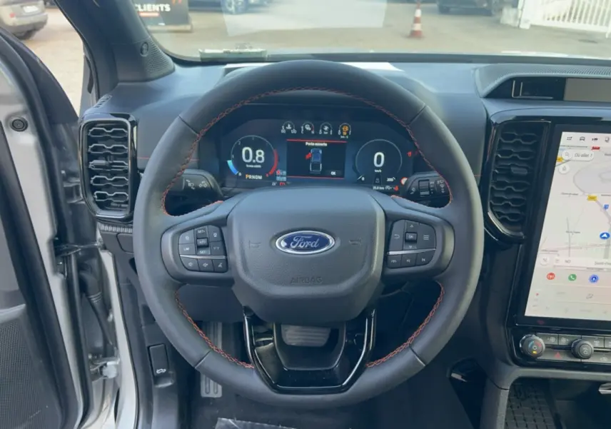Vue centrée sur le volant noir avec coutures rouges du Ford Ranger Wildtrak 2026, tableau de bord digital et écran tactile GPS visible.