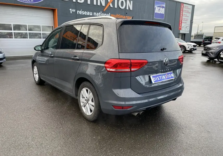 Vue 3/4 arrière droite d'un Volkswagen Touran gris dauphin avec vitres arrière surteintées, stationné devant un garage multimarque.