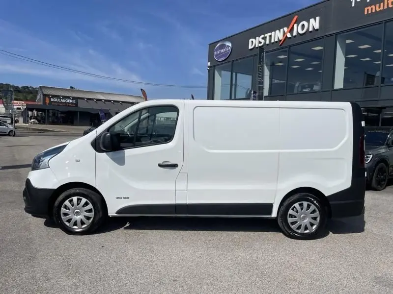 Vue de profil côté gauche d’un Renault Trafic Fourgon blanc L1H1 DCI 125 cv, stationné devant un bâtiment commercial.