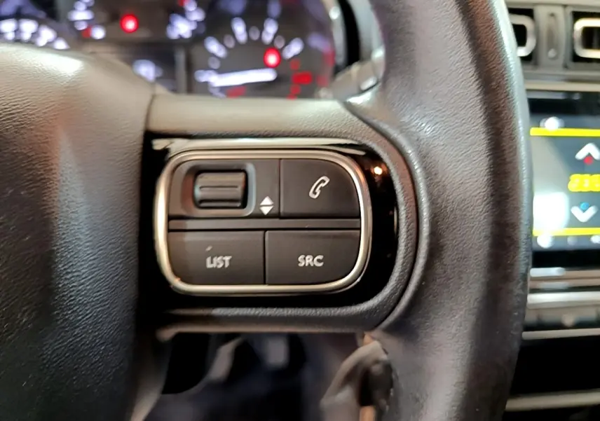 Gros plan sur les commandes gauche du volant cuir de la Citroën C3 blanche, avec tableau de bord et écran tactile en arrière-plan.