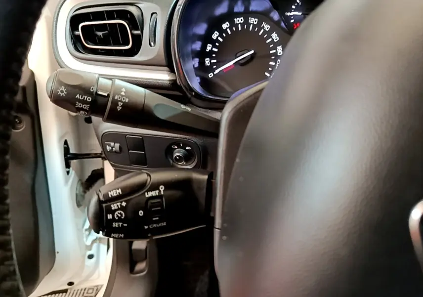 Vue intérieure côté conducteur de la Citroën C3 blanche, volant cuir et commandes au volant visibles avec tableau de bord éclairé.