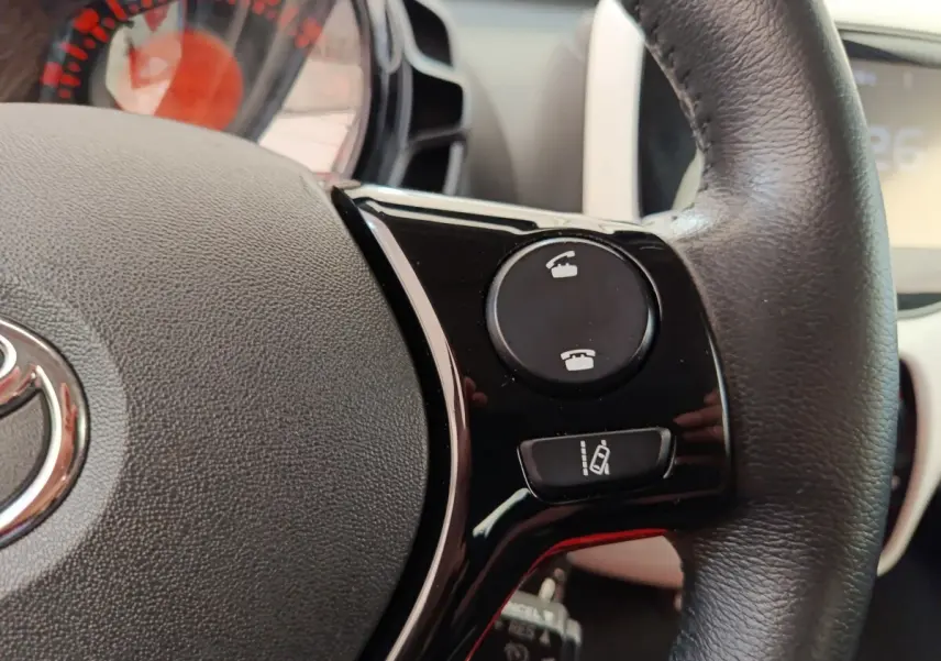 Gros plan sur le volant cuir noir de la Toyota Aygo blanc 2016, avec commandes téléphoniques et aide au maintien de voie.