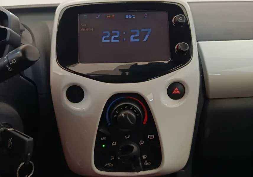 Vue rapprochée du tableau de bord blanc de la Toyota Aygo 2016, avec écran digital affichant l'heure et commandes de climatisation.