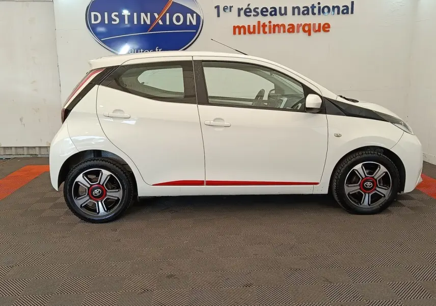 Profil latéral droit de la Toyota Aygo blanche 2016 avec jantes noires et rouges et détails rouges sur les bas de caisse.