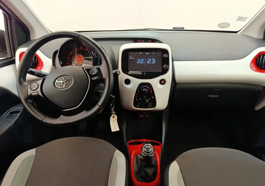 Vue intérieure frontale de la Toyota Aygo blanche, volant cuir, console centrale avec écran tactile et levier de vitesse rouge.
