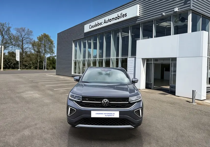Volkswagen T-Cross gris vu de face devant un showroom avec calandre noire et feux LED allumés.