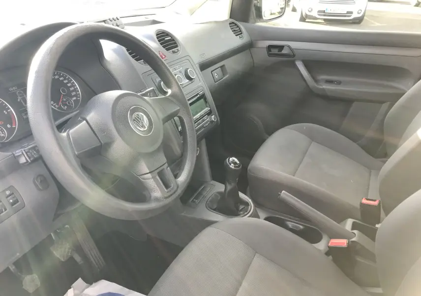 Intérieur avant du Volkswagen Caddy Maxi Van blanc 2015, vue côté conducteur montrant volant, tableau de bord et sièges tissu noir.