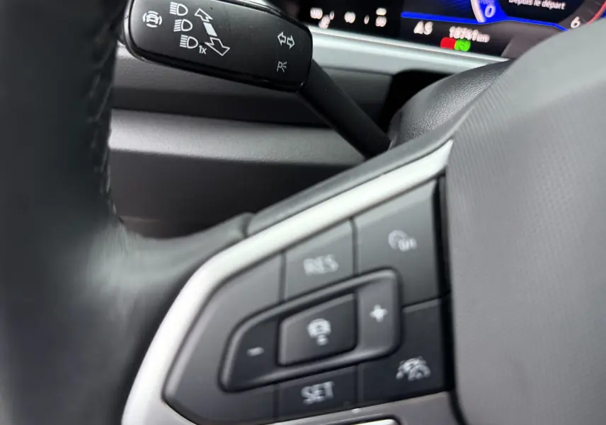 Gros plan sur les commandes au volant côté gauche du Volkswagen T-Roc 2025, avec tableau de bord numérique en arrière-plan.