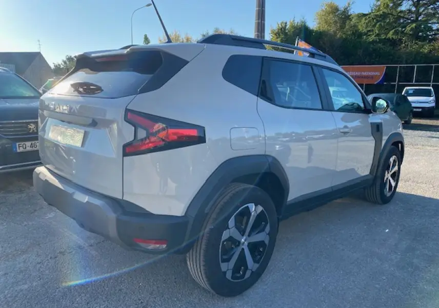 Vue 3/4 arrière droite du Dacia Duster 2025 beige Sandstone avec jantes alliage et barres de toit noires.