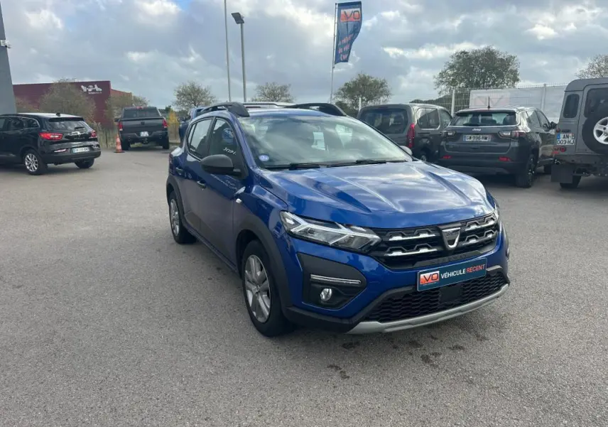 Vue 3/4 avant d'une Dacia Sandero Stepway bleu foncé avec barres de toit, garée sur un parking extérieur.