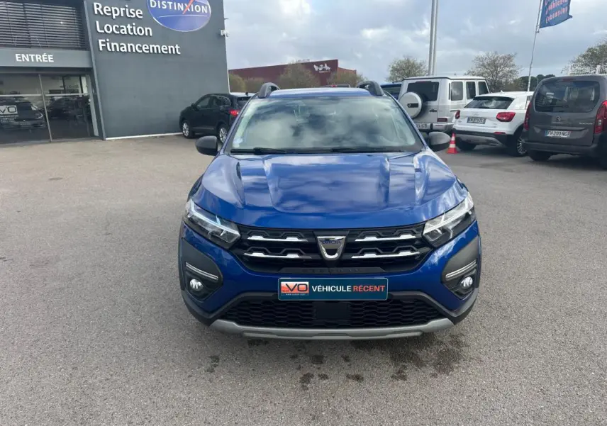 Vue frontale d'une Dacia Sandero Stepway bleu foncé avec barres de toit et feux allumés sur parking.