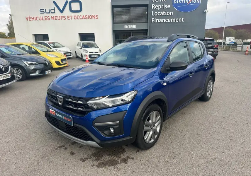 Vue 3/4 avant d'une Dacia Sandero Stepway bleu foncé avec barres de toit et phares allumés sur parking.