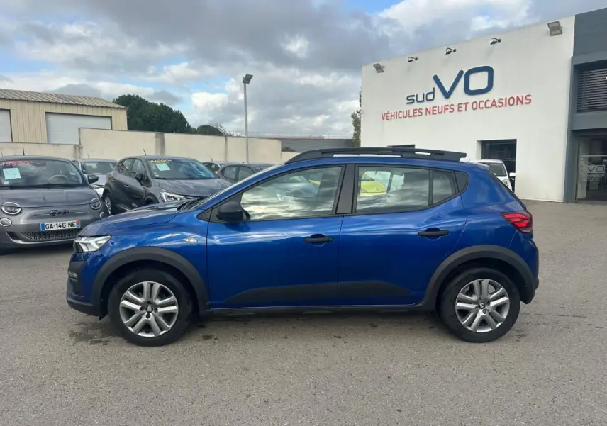 Profil côté gauche d'une Dacia Sandero Stepway bleu foncé avec barres de toit et vitres surteintées, stationnée en extérieur.