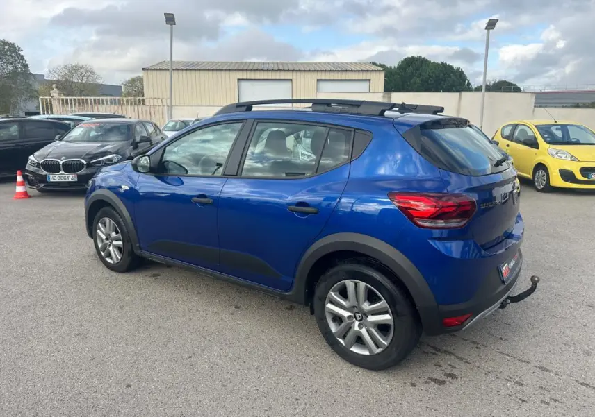 Vue 3/4 arrière gauche d'une Dacia Sandero Stepway bleu foncé avec barres de toit et attelage visible.