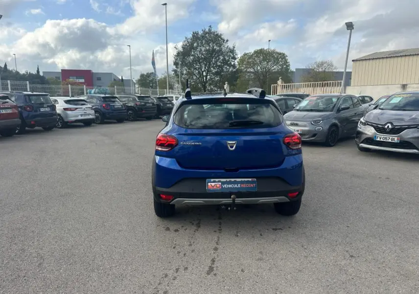 Vue arrière d'une Dacia Sandero Stepway bleu foncé avec barres de toit et attelage sur un parking.