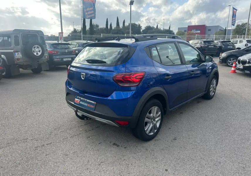 Vue 3/4 arrière droite d'une Dacia Sandero Stepway bleu foncé avec barres de toit et vitres surteintées dans un parking.