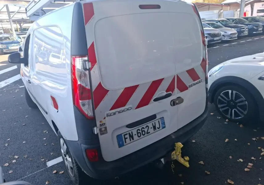 Vue 3/4 arrière droite d’un Renault Kangoo blanc avec bandes rouges réfléchissantes sur les portes arrière, stationné sur un sol gravillonné.