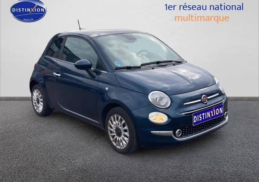 Vue 3/4 avant gauche d'une Fiat 500 bleu foncé avec jantes alu et phares allumés sur fond neutre.