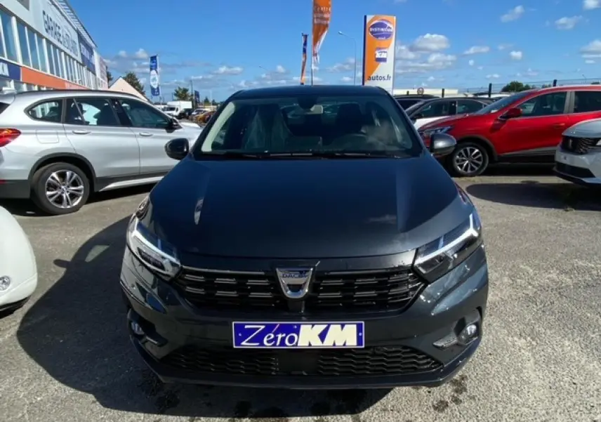 Vue frontale d'une Dacia Logan gris foncé 2022 avec calandre noire et plaque "Zero KM" sur un parking extérieur.