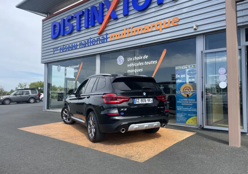 BMW X3 noir vue 3/4 arrière droit, stationné devant une concession avec vitres réfléchissantes et enseigne colorée.