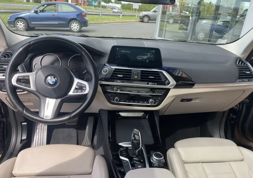 Vue intérieure du tableau de bord beige et noir d’un BMW X3 G01 2021, avec volant multifonction et écran central allumé.