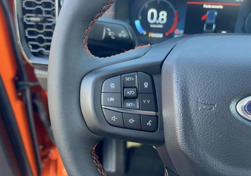 Gros plan sur le volant multifonctions noir avec surpiqûres orange du Ford Ranger Wildtrak Ignite orange, tableau de bord digital en arrière-plan.