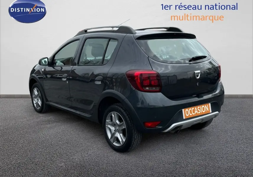 Vue 3/4 arrière droite d'une Dacia Sandero Stepway 0.9 TCe gris foncé de 2016 avec barres de toit et jantes alliage.